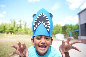 Laughing child wearing a blue paper shark hat and making a scary face outdoors