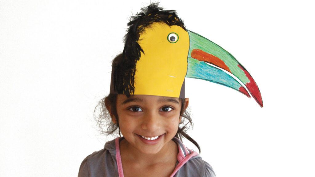 Smiling girl wearing a colorful paper toucan headband with green and red bill and feathers