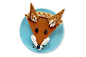 Slice of pumpkin pie decorated to look like a fox face with whipped cream cheeks and olive eyes