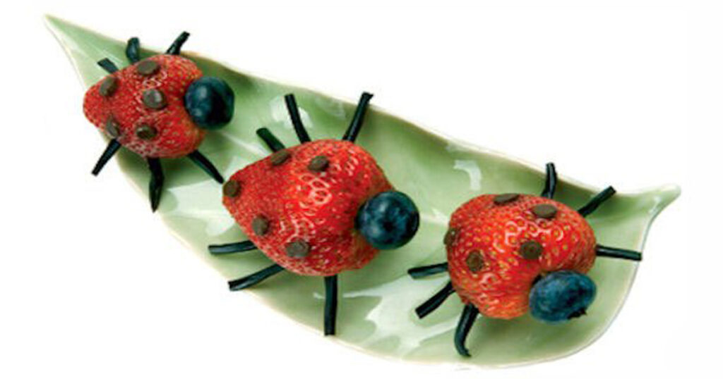 Three strawberry ladybugs with blueberry heads and licorice legs on a green leaf plate