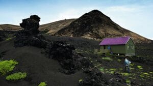Small green research hut on black volcanic lava landscape of Surtsey with scientist walking