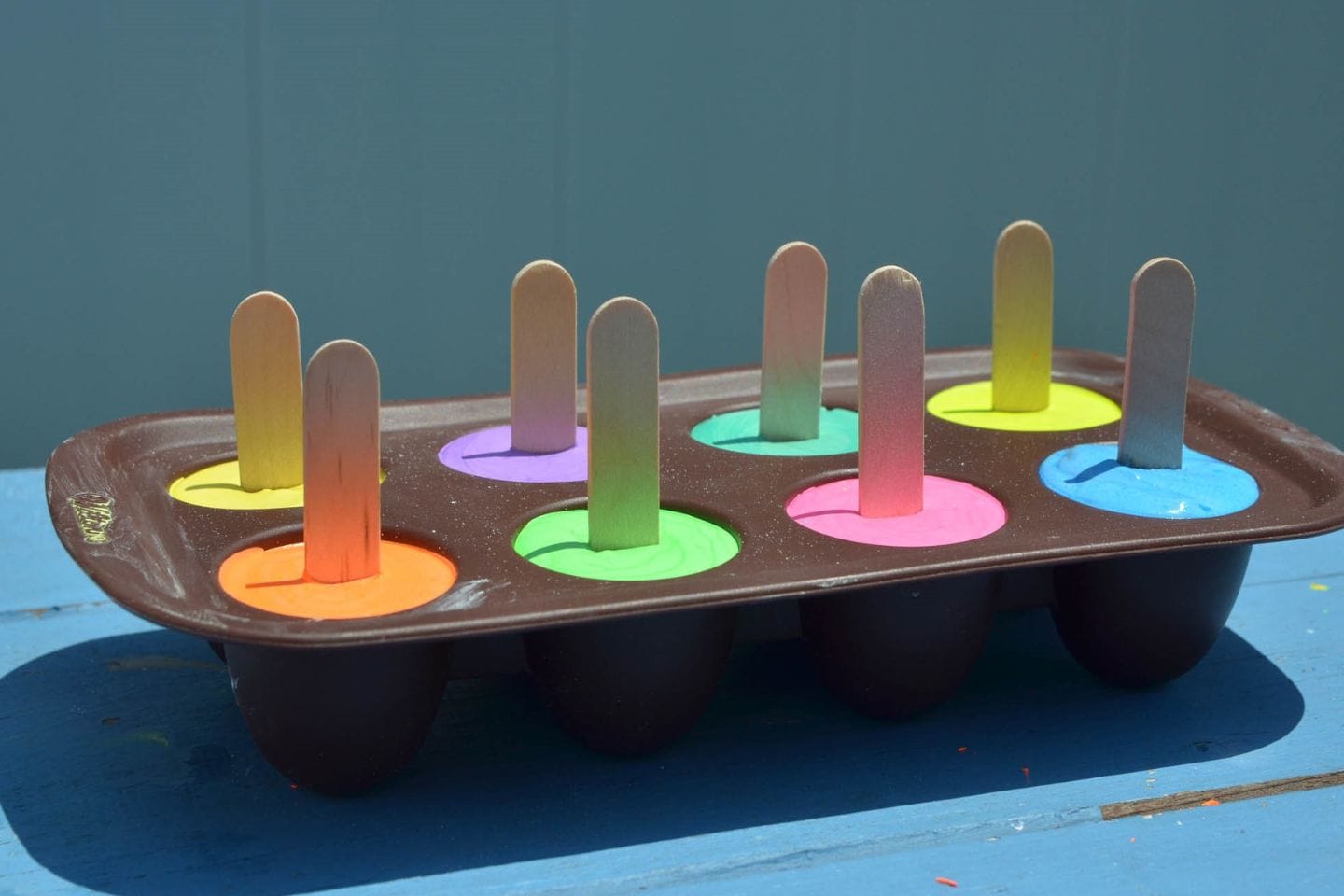 Colorful homemade sidewalk chalk pops in a silicone popsicle mold with wooden craft sticks, drying outdoors in the sunshine.