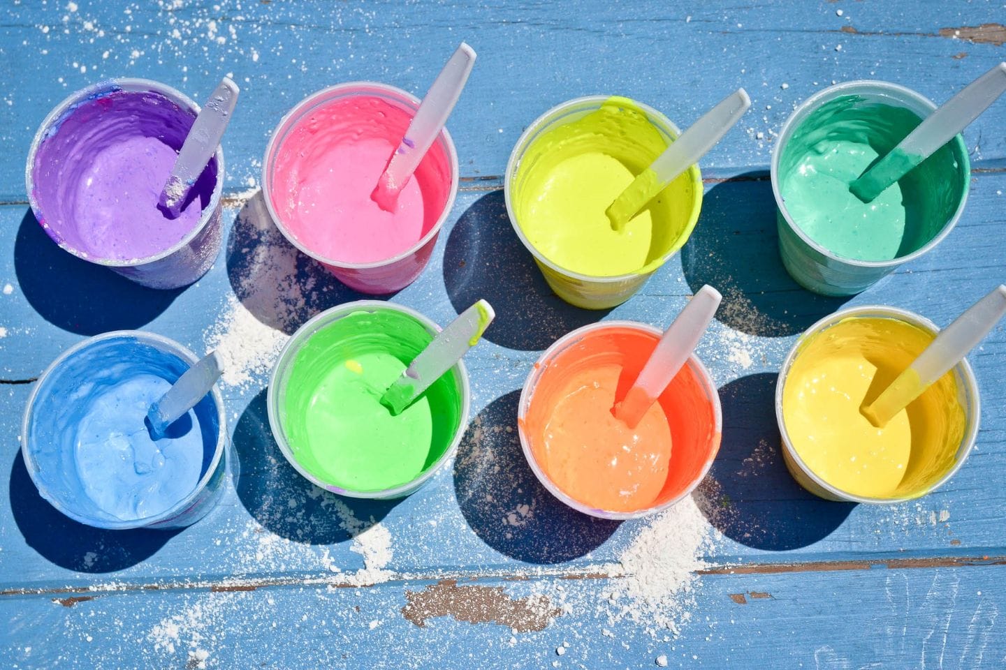Disposable cups filled with colorful plaster and tempera paint mixtures for making homemade sidewalk chalk, each with a stirring stick on a blue table.