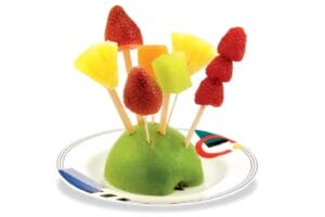 half of a sliced green apple base with skewers of summer fruit on a white plate