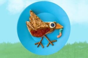Toast shaped like a robin bird with peanut butter wings, jelly breast, pretzel legs, and a gummy worm beak